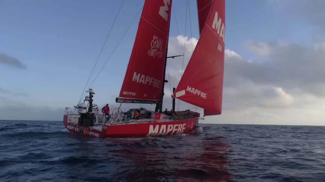 Drone shots: Distant shot with sunrise; very low-altitude shot overtaking boat from the starboard quarter. Shots on board of slatting sails, instruments with low-wind/low-speed readings. At night, Joan and Xabi talking in Spanish at nav station about a position report. Then, in the dark, we hear Joan (?) relaying the position report in English to Rob (?), talking about how they're doing vs. Vestas (ahead of them) and the peleton (behind them).