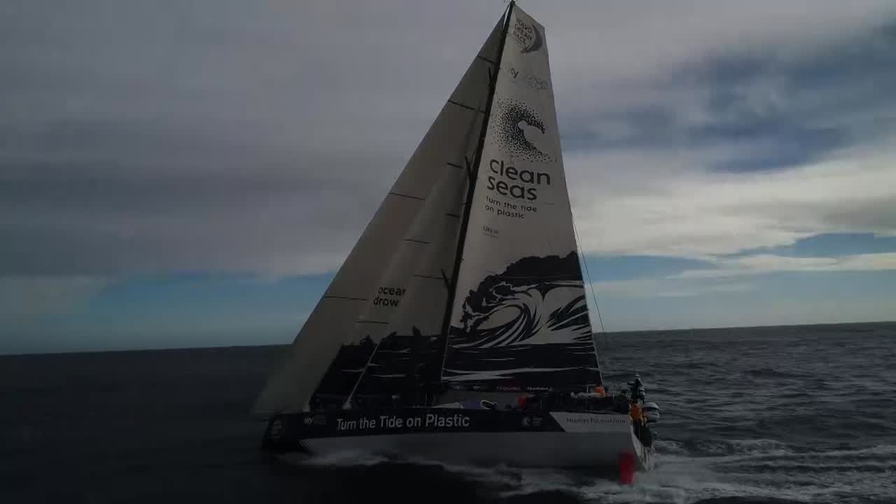 Drone shots of TTToP sailing on port gybe, triple-heading with the MH0 (I think?) in about 15 knots of wind. Later, the drone looks down from above, and the J2 has been furled. Below, Nicolas sits at the nav station looking at routing software. We see him by the wheel, explaining to Liz, who's steering. Liz looks over the side at the area of the rudder. Someone (Elodie?) grinds. Wake through wide-angle lens. Wide-angle shot from behind as Lucas steers, surfing. He turns around briefly, taking both hands off the wheel to make dual shaka signs and stick his tongue out. Francesca, trimming the main, chuckles. They surf in increased wind. Bleddyn coils in the pit. A wave douses the pit. Later, it's almost dark, we see out from the cabin hatch as water washes into the cockpit.