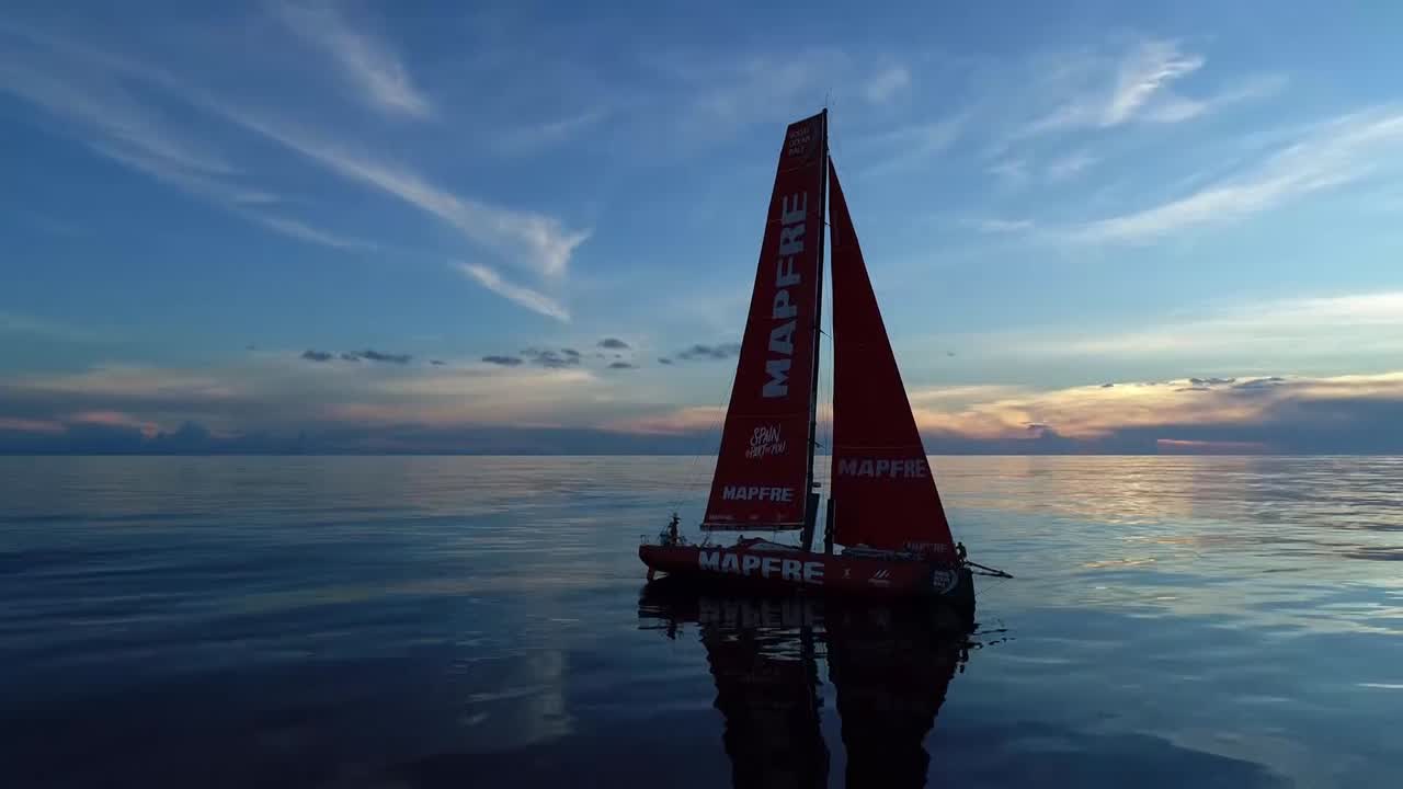 We see Ugo launch the drone, which flies away. MAPFRE drifts on a glassy ocean. Very low-altitude drone shots that circle the boat, first farther away and then close.At one point we see a distant competitor ahead and to leeward. We see Sophie's silhouette on the bow as they hoist the J1 (or maybe it's just that the J1 is set with a loose halyard for drifting/windseeking conditions).